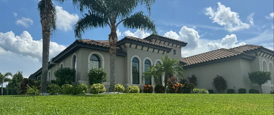 Luxurious house with Mediterranean architectural features, surrounded by a well-manicured lawn and palm trees. The sky is blue with scattered clouds.